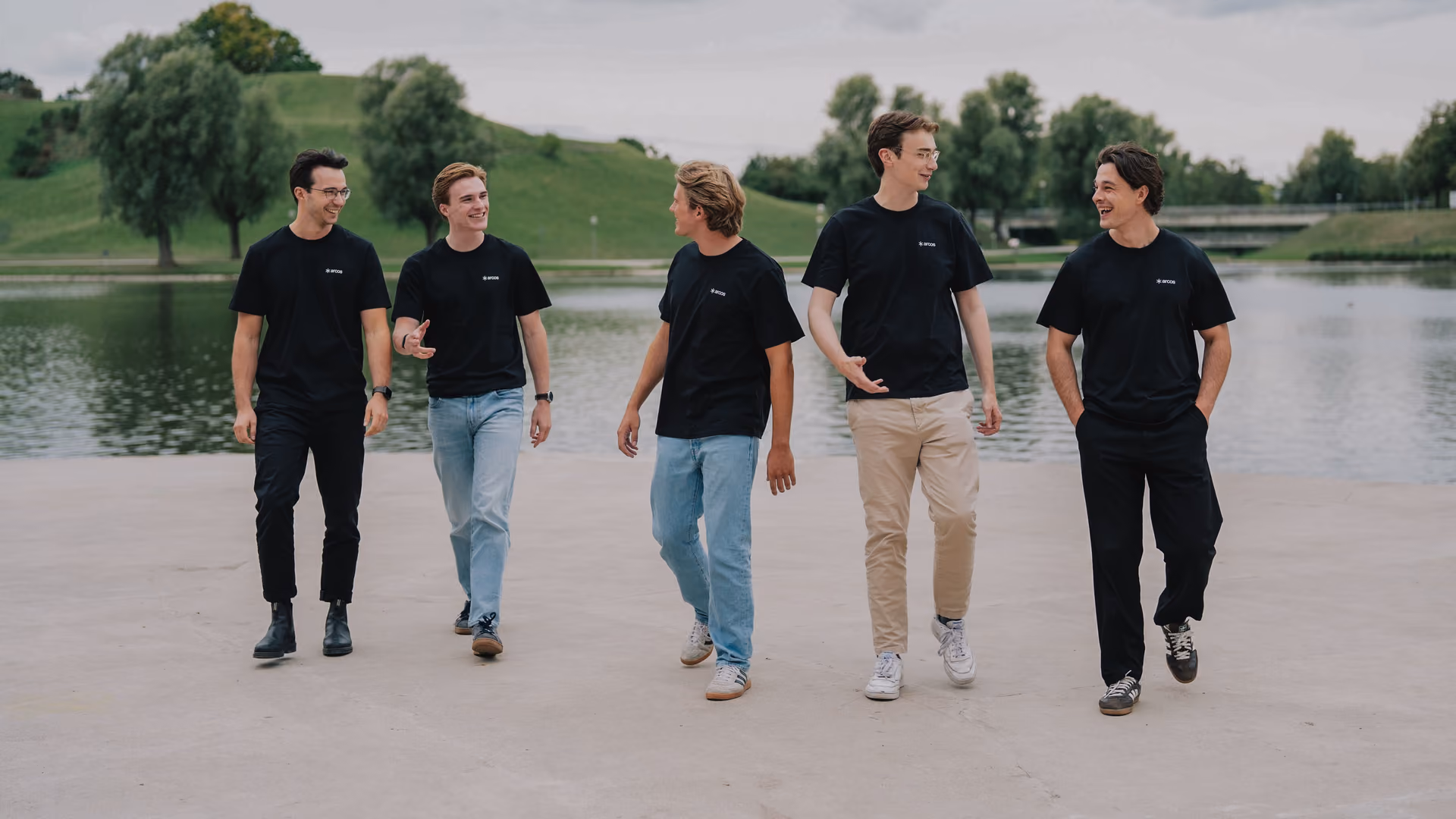 Five people in black t-shirts walking side by side beside a lake.
