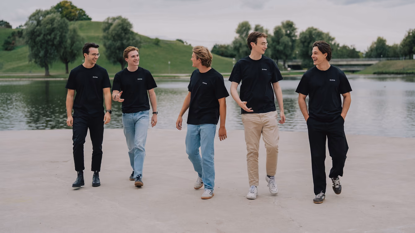 Five people in black t-shirts walking side by side beside a lake.
