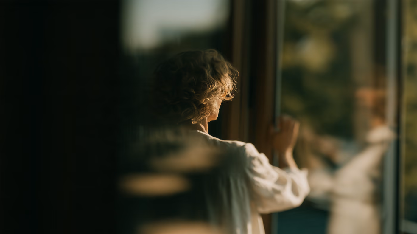 A woman at a window, one hand on the frame, looking out.
