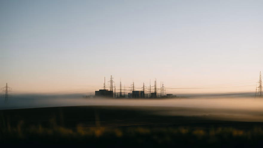 A high-voltage substation with transmission pylons standing in morning fog above a field.