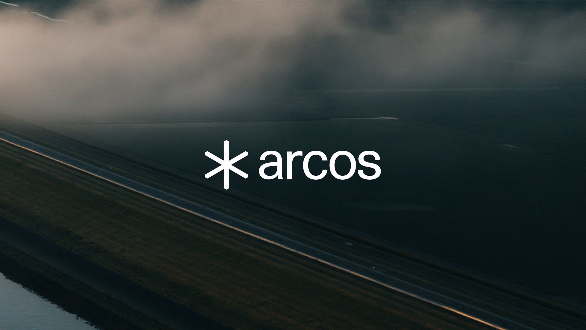 The Arcos star and wordmark over an aerial view of a road running past a dark, flat expanse under low cloud.